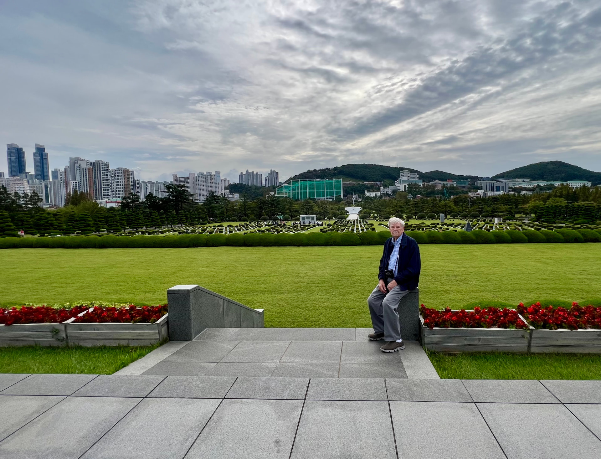 Laura's father sitting quietly at the Korean War memorial in Busan, with the modern city skyline behind him