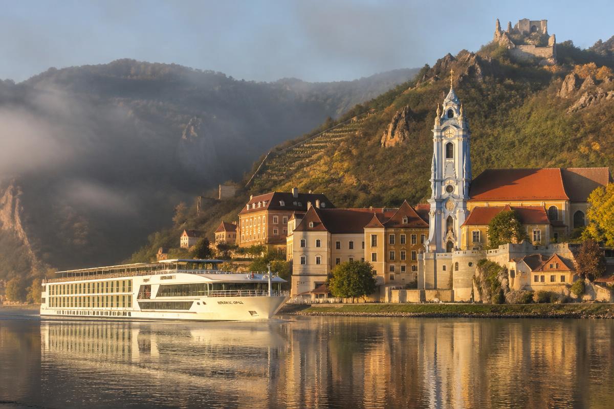 Elegant river cruise ship passing a historic European village with vineyards and a baroque church tower